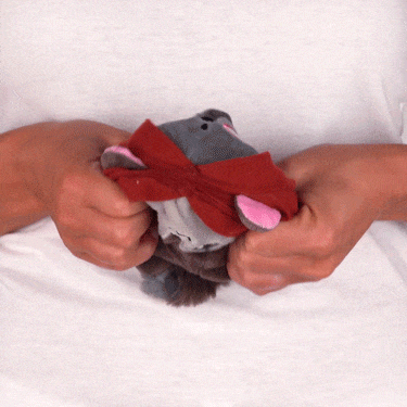 A person's hands untangling a Casting Shadows: Nuzzle Thornwood & Nuzzle the Savage Reversible Plushie rat's tail from a red ribbon on a white background by Unstable Games.