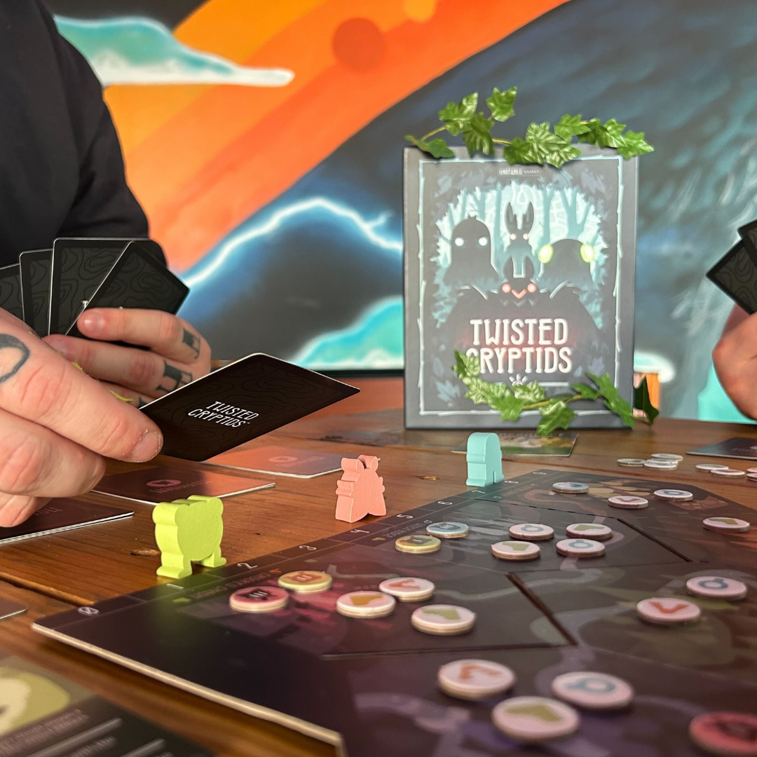Two people play Twisted Cryptids: Base Game by Unstable Games on a wooden table, holding cards with game pieces on the board; the game box and a vibrant mural are visible in the background.