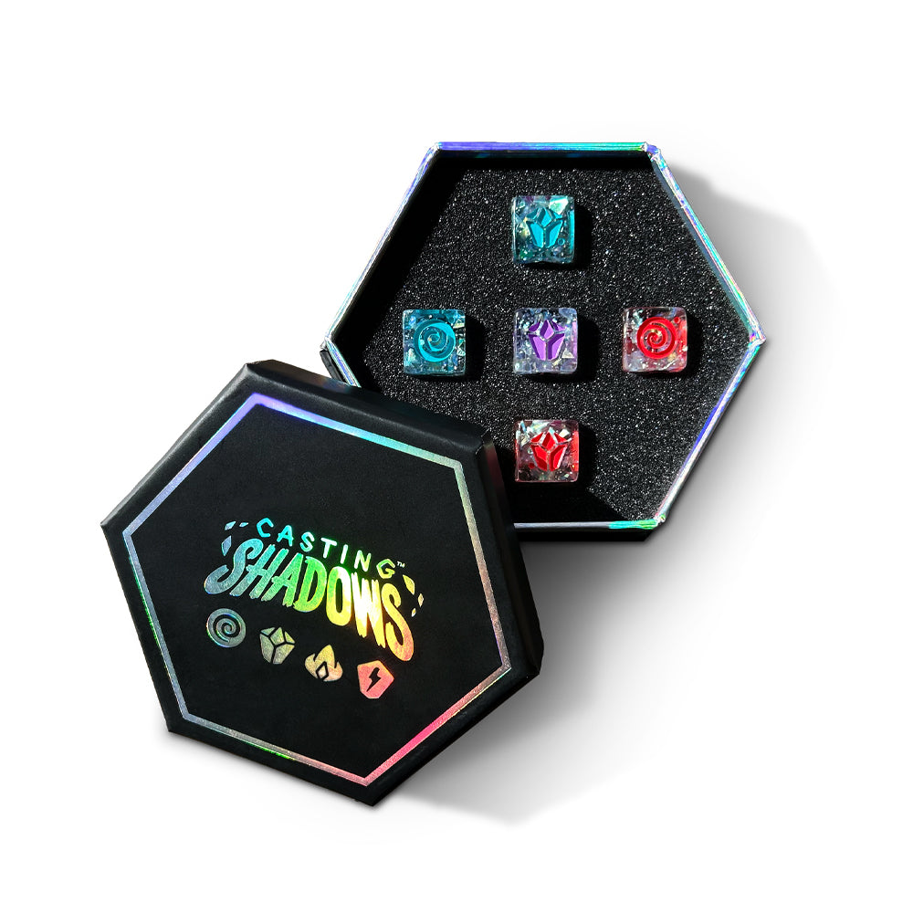 A hexagonal black box containing six colorful, Casting Shadows: Sparkle Core Resource dice, with the box lid featuring the text "Casting Shadows" beside dice icons by Unstable Games.