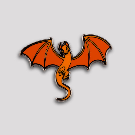 Dragon Skies Pin by Unstable Games: orange dragon-shaped pin with wings outstretched, designed with bold outlines and highlighted details on a gray background.