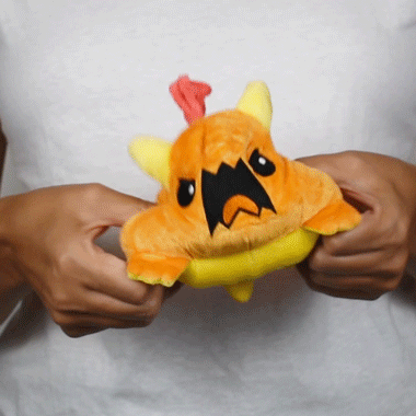 A person holding and slightly squeezing a small, Tic Tac K.O. Dragons vs. Unicorns: Tantrum & Book Wyrm reversible plushie with a sad face and chicken-like features from Unstable Games.