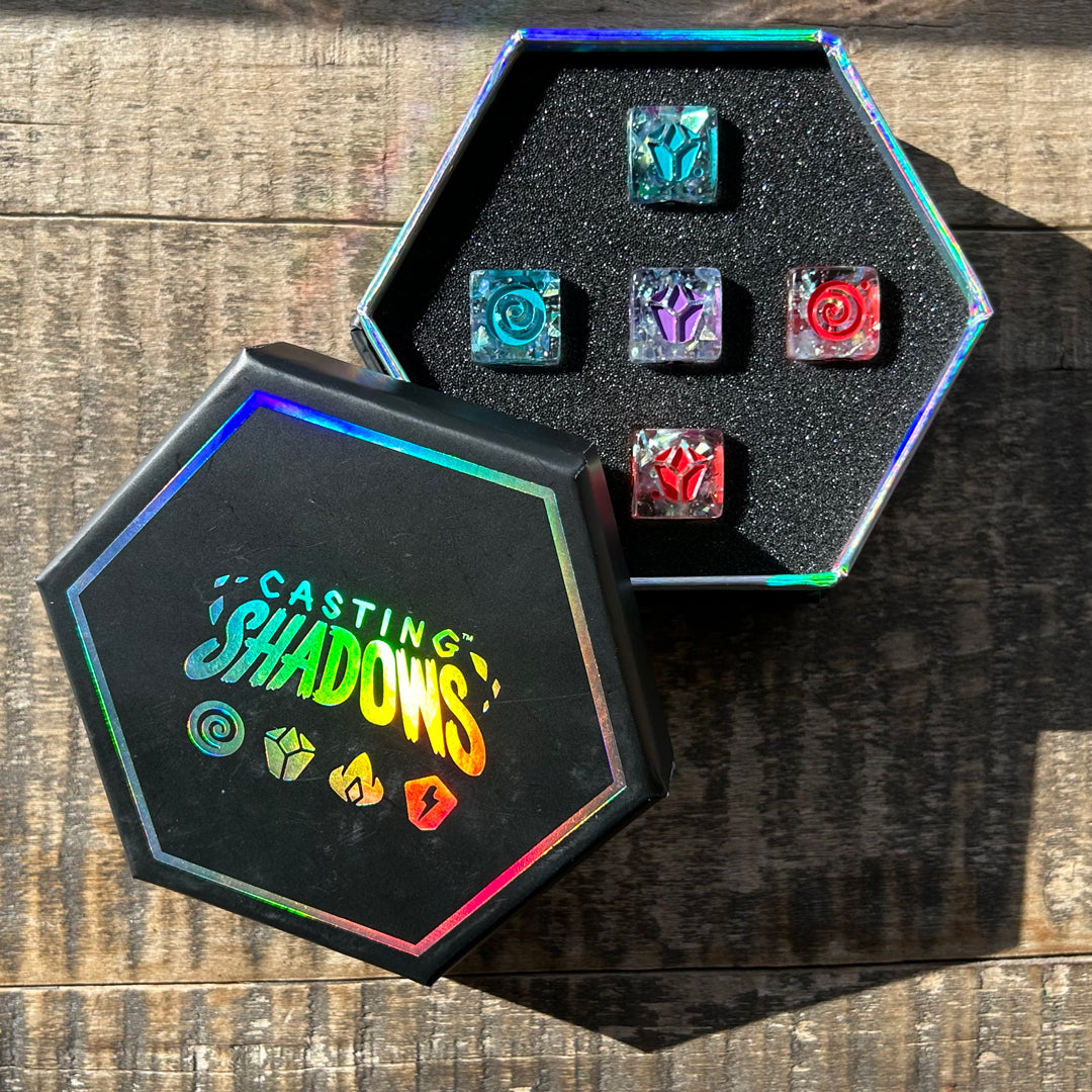 A hex-shaped box with the text "Casting Shadows: Sparkle Core Resource Dice Set" on the lid, containing six colorful, polyhedral dice set in a foam insert, on a wooden surface. Brand Name: Unstable Games
