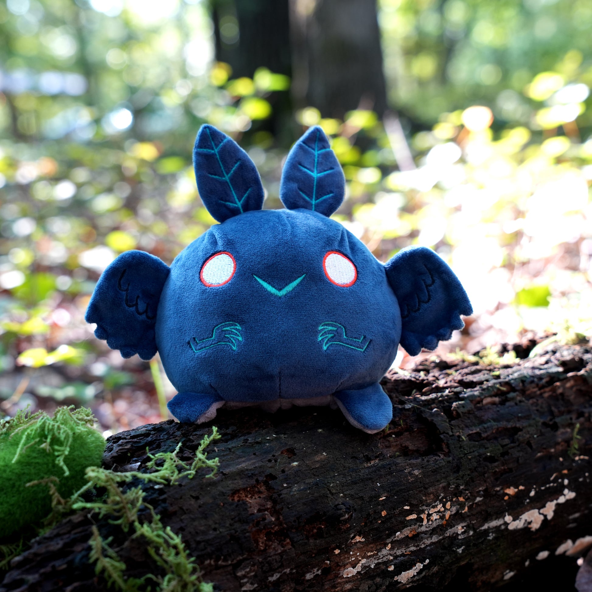 Twisted Cryptids: Mothman 4" Reversible Plushie happy side sitting on a mossy log in a forest