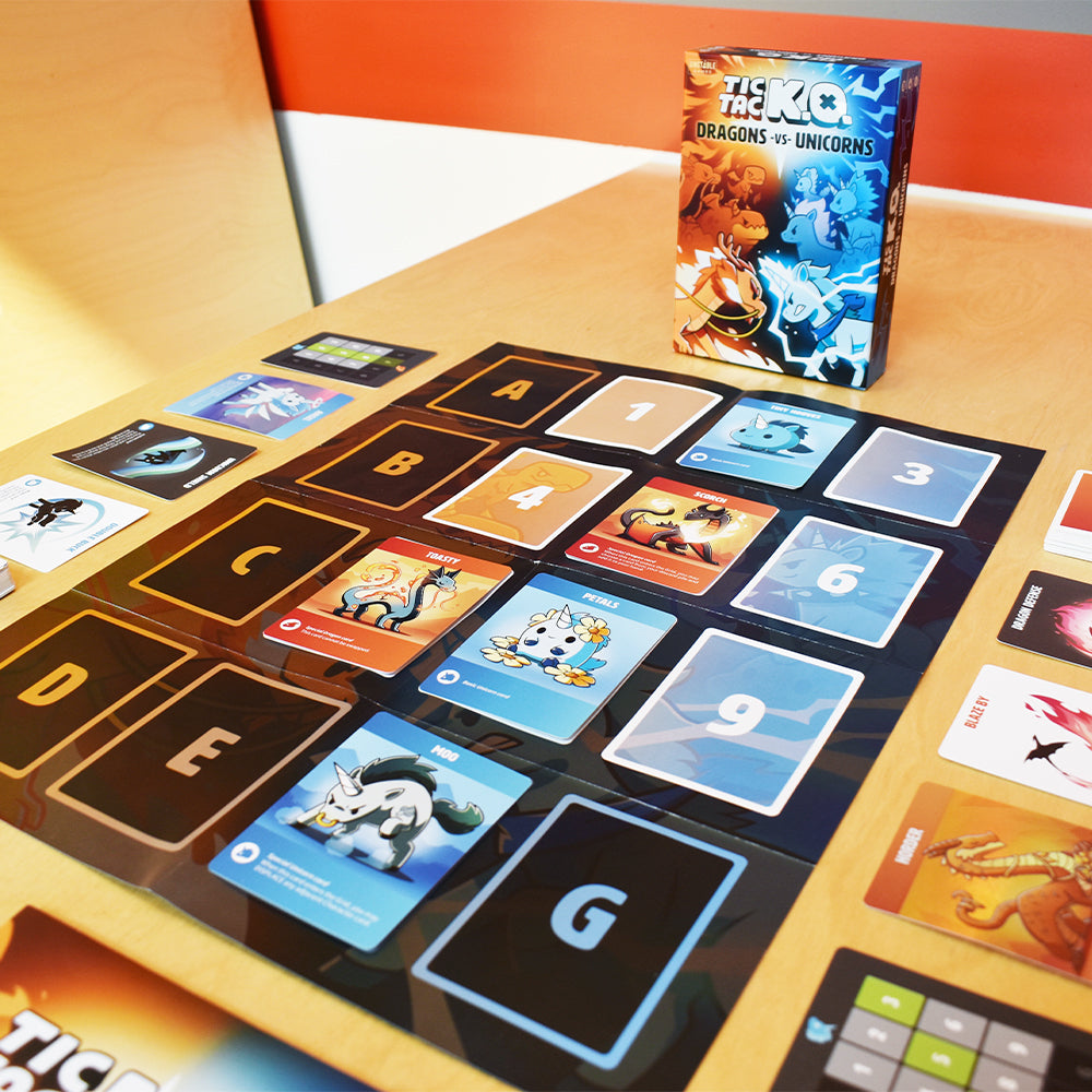 A card game titled "Tic Tac K.O. Dragons vs. Unicorns: Base Game" by Unstable Games is displayed on a table with various illustrated cards laid out in a grid pattern.